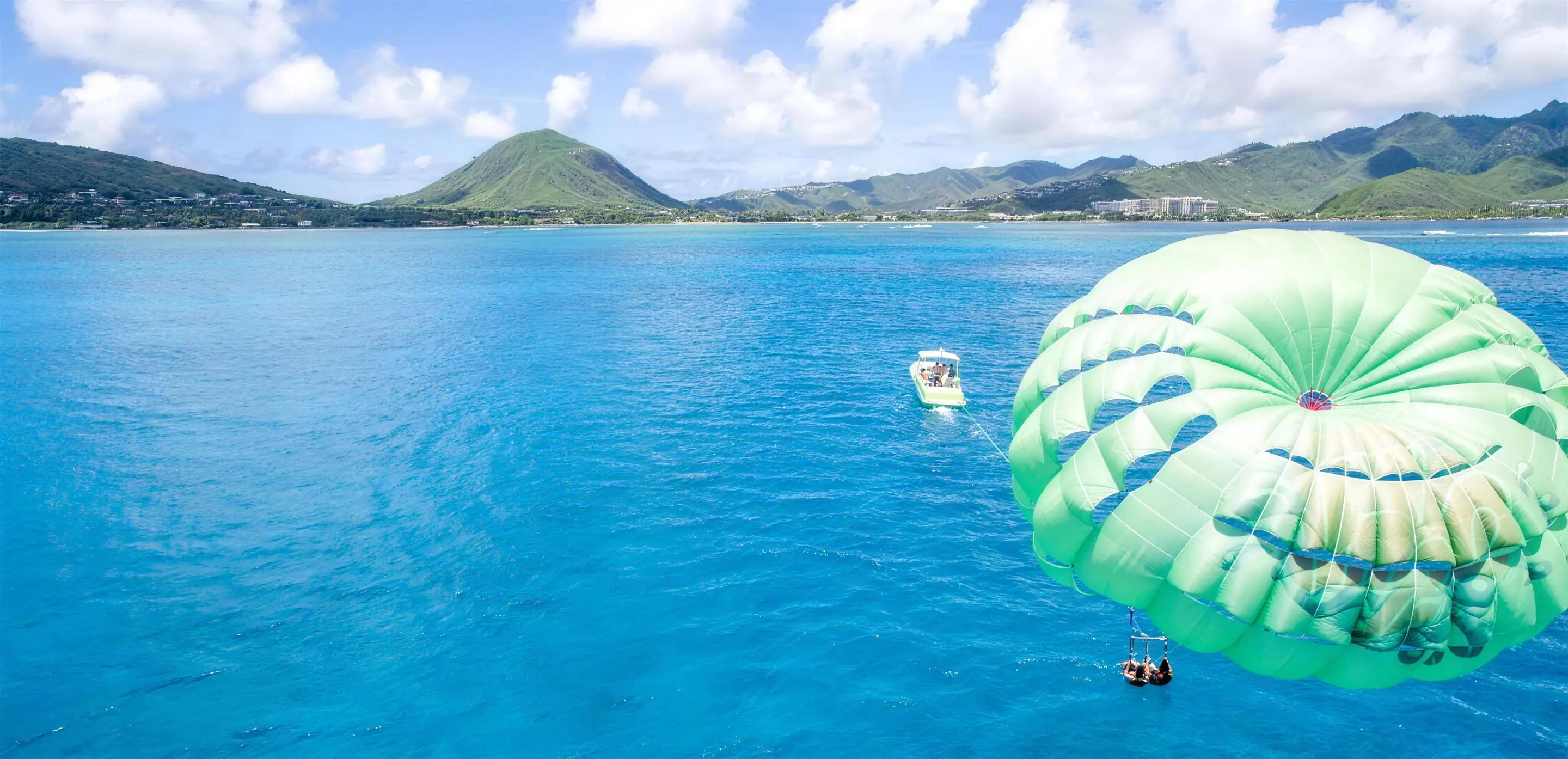 Two people under a lime-green parasail towed by a boat, with Koko Head behind