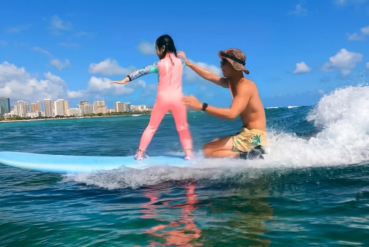A child standing on a board as an instructor holds it from behind, with Waikiki buildings in the background