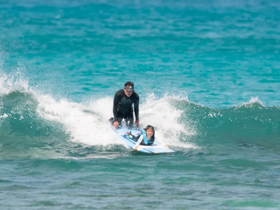 A smiling child lying on a board on a wave as an instructor kneels behind to hold it